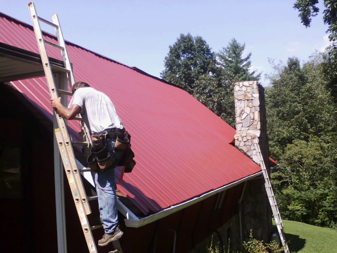 Roofer on a ladder beside a red metal roof and stone chimney — Soffit & Fascia Repair by Roofing Brothers in Hotchkiss